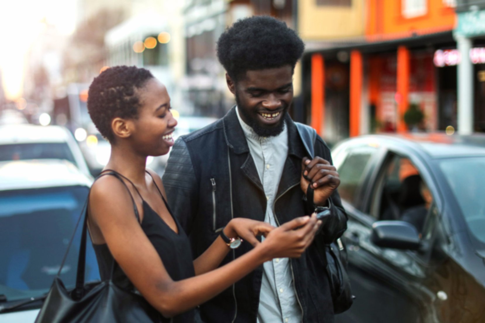 Couple walking through traffic, they're both laughing as the woman is showing the man something on her phone.